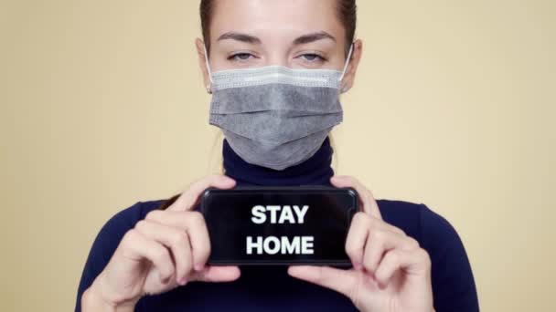 Retrato de mujer joven en máscara médica muestra teléfono con inscripción estancia en casa — Vídeo de stock