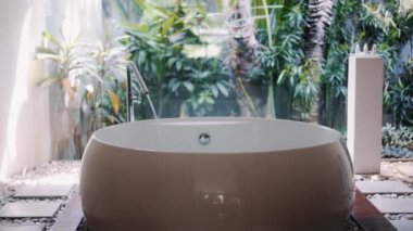 Yuvarlak küvete su doluyor, lüks banyo, arka planda tropik yeşillik.