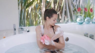 Bikinili bir kadın vücudu havluyla yıkıyor ve yuvarlak bir küvette banyo yapıyor.