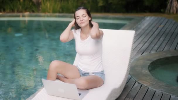 Woman works on laptop sitting on sunbed near pool, enjoying sunny day — Stock Video