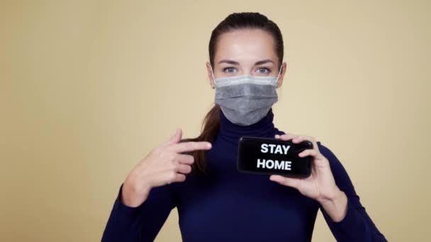 Porträtt av kvinna i medicinsk mask visar på telefon med inskription stanna hemma — Stockvideo