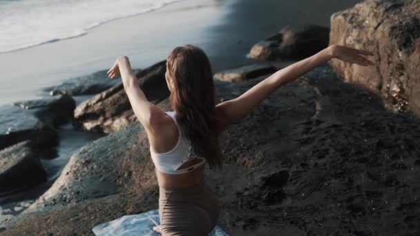 Вид ззаду, дівчина робить йогу тягнеться на килимок, чорний піщаний пляж біля океану — стокове відео