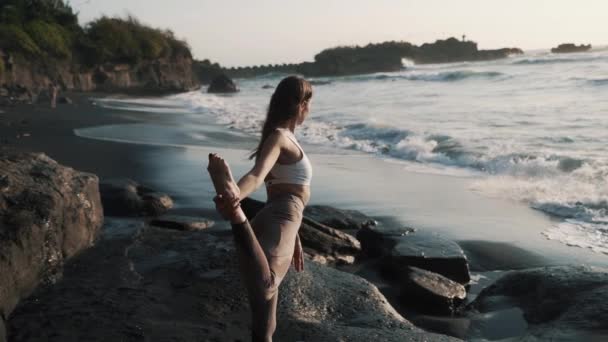 Visão traseira, menina fazendo ioga alongamento, praia vulcânica de areia preta perto do oceano — Vídeo de Stock