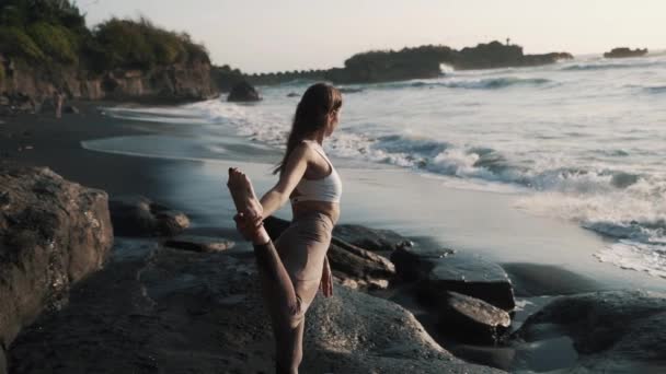 Vista trasera, chica haciendo yoga estiramiento, playa volcánica de arena negra cerca del océano — Vídeos de Stock