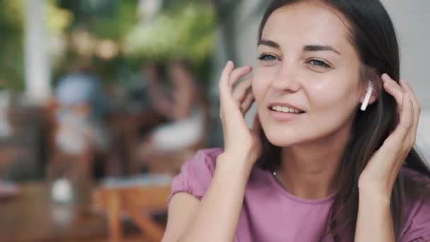 Молодая женщина надевает беспроводные наушники, слушает музыку и танцует в кафе — стоковое видео