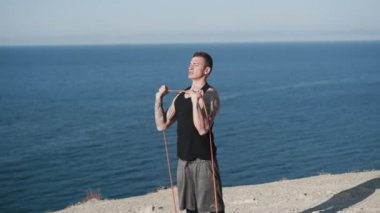 Dövmeli bir adam spor yapıyor. Açık havada, deniz manzaralı lastik bantlarla.