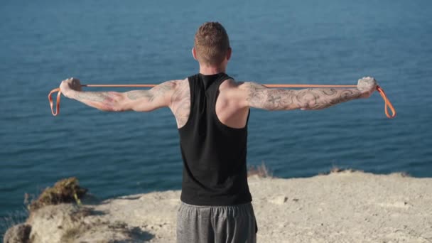 Backside view of muscular tattooed sportsman trains with elastic bands outdoor — Stock Video