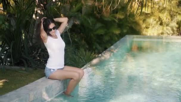Девушка в солнечных очках и летней одежде сидит на краю бассейна, держит ноги в воде — стоковое видео