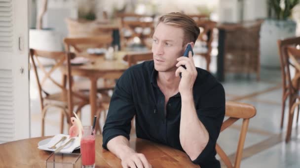 Retrato de hombre de negocios con camisa negra sentado en la cafetería y hablando por teléfono — Vídeos de Stock