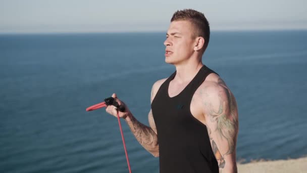 Jovem tatuado faz exercícios com bandas elásticas ao nascer do sol com vista para o mar — Vídeo de Stock