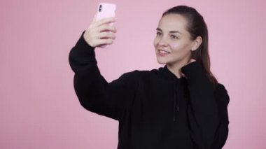 Kadın gülümsüyor ve cep telefonuyla özçekim yapıyor, pembe arka planda izole edilmiş.