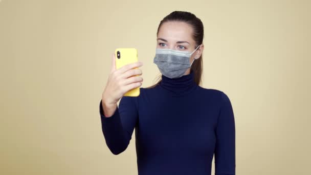 Kvinna i medicinsk mask gör selfie på mobiltelefon, isolerad på beige bakgrund — Stockvideo