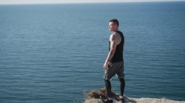Dövmeli adam dağın tepesinde durur, arkasını döner, kameraya bakar, deniz manzarası