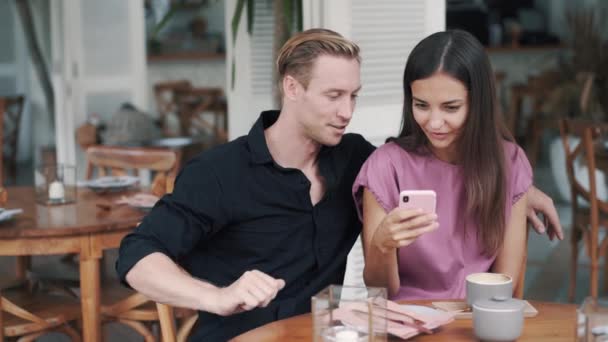 Giovane uomo e donna seduti nel caffè, guardando smartphone, ridendo e parlando — Video Stock