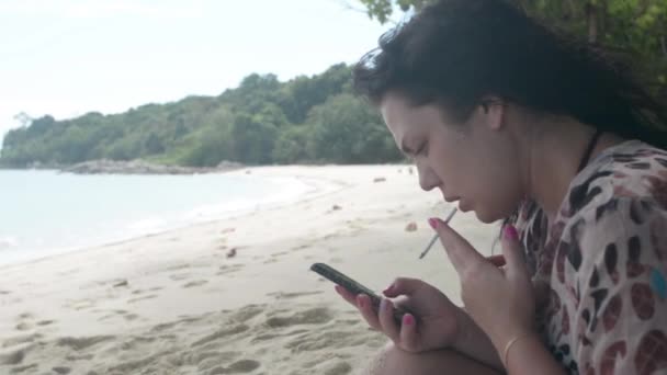 女孩坐在一个野沙滩上 抽着烟 看着智能手机 — 图库视频影像