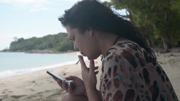 女孩坐在一个野沙滩上 抽着烟 看着智能手机 — 图库视频影像