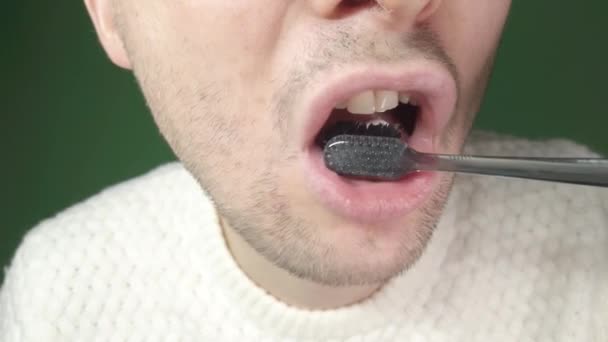 Fiatal férfi fogat mos egy fekete fogkefével, közelkép zöld háttérrel — Stock videók