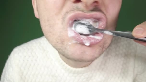Fiatal férfi fogat mos egy fekete fogkefével, közelkép zöld háttérrel — Stock videók