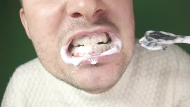 Young man brushing his teeth with a black toothbrush, close-up on a green background — Stock Video