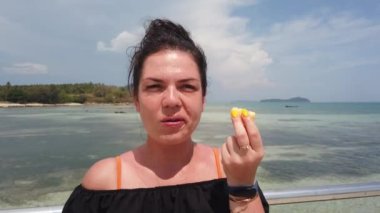 Kız gök mavisi denize karşı iskelede duruyor ve sulu bir ananas yiyor.