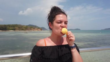 Kız gök mavisi denize karşı iskelede duruyor ve sulu bir ananas yiyor.