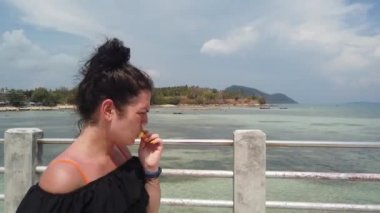 Kız gök mavisi denize karşı iskelede duruyor ve sulu bir ananas yiyor.