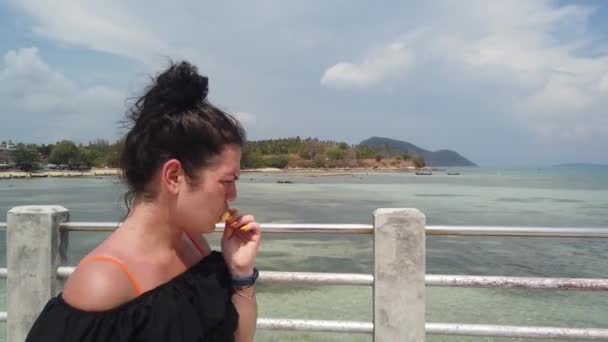 少女は紺碧の海に対して桟橋に立って 小さなジューシーなパイナップルを食べる — ストック動画