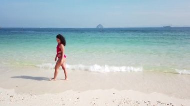 Esmer kız, ıssız bir adanın kumsalında masmavi suyla yürüyor.