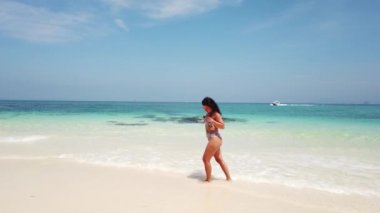 Esmer kız, ıssız bir adanın kumsalında masmavi suyla yürüyor.