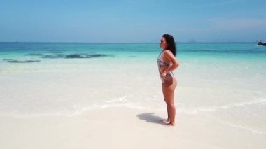 Esmer kız, ıssız bir adanın kumsalında masmavi suyla yürüyor.