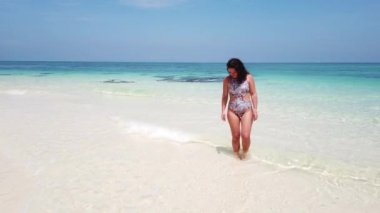 Esmer kız, ıssız bir adanın kumsalında masmavi suyla yürüyor.