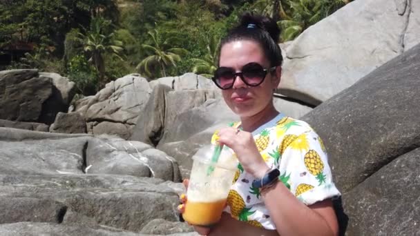 熱帯の背景にある大きな石の上に座っているビーチで新鮮な果物を飲む女の子 — ストック動画
