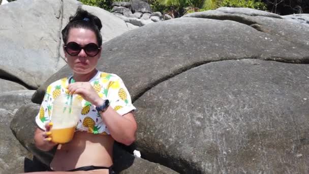 熱帯の背景にある大きな石の上に座っているビーチで新鮮な果物を飲む女の子 — ストック動画