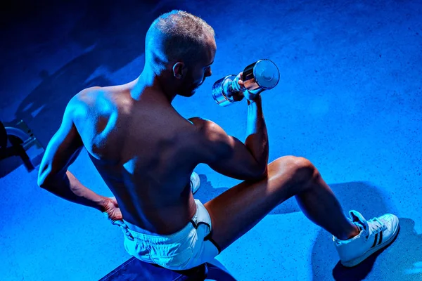 Homem sem camisa sentado no banco e levantando halteres — Fotografia de Stock