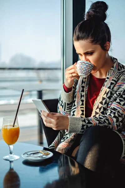 Girl drinking coffee and using phone by the window — Stock Photo, Image