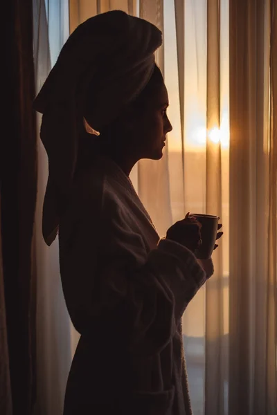 Woman in bathrobe looking through window and holding cup — Stock Photo, Image