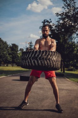 Sakallı sporcu parkta lastik kaldırıyor.