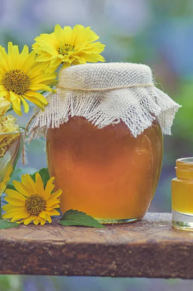 Frascos de miel, panal de abeja sobre mesa de madera con flores de fondo —  Fotos de Stock