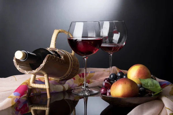 Dois copos de vinho tinto, garrafa em suporte de palha e placa de madeira com vinhas e maçãs situadas na mesa de espelho pretocoberta por pano rosado — Fotografia de Stock