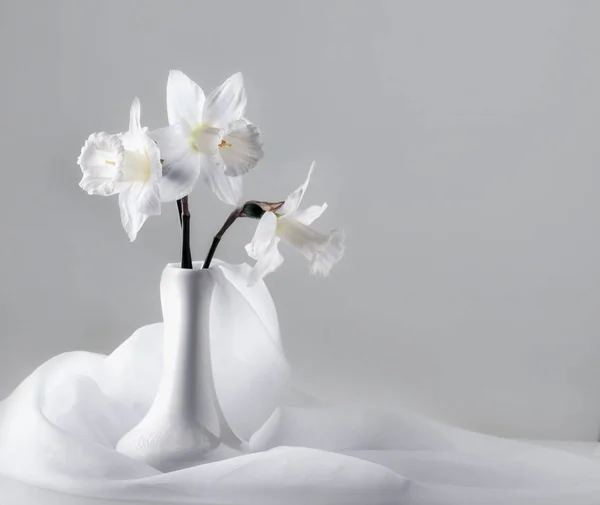 Bouquet of white narcissus flowers in a vase surrounded by white piece of veil fabric on light grey background. Wedding invitation card or present card. — Stock Photo, Image