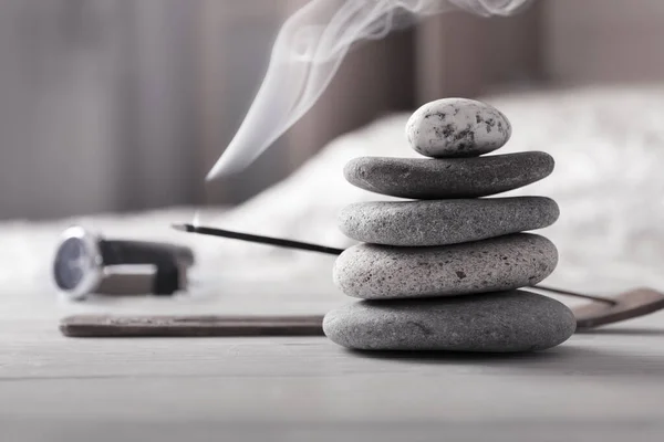 Evening meditation at home. Pyramid of zen stones, burning incense stick next to watch lying on white wooden table in front of bed. — Stock Photo, Image