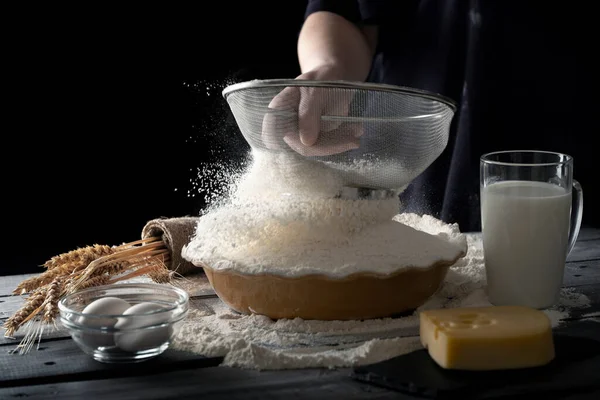 Wanita menyaring tepung dalam mangkuk di atas meja kayu. Latar belakang gelap. Bahan adonan. Stok Lukisan  