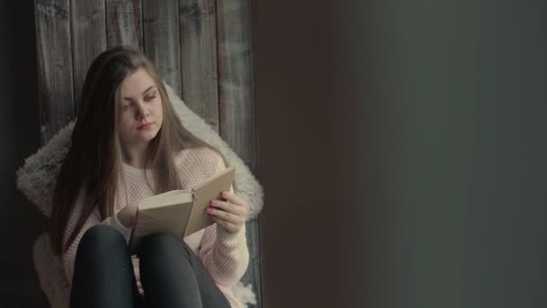 Beautiful young woman sitting on window and read a book turning pages — Stock Video