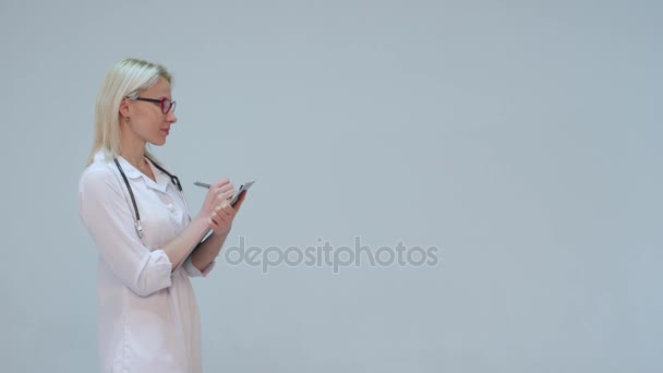Dokter in een witte badjas en stethoscoop glimlacht, records de instrumenten — Stockvideo