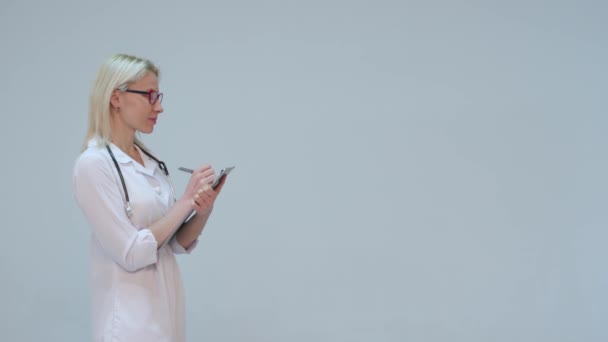 Блондинка Доктор пишет на планшете, улыбаясь на сером фоне — стоковое видео