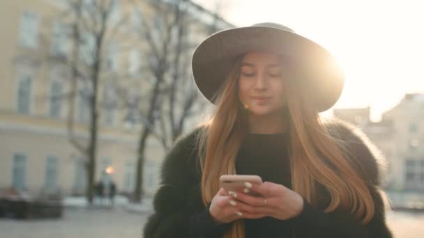 Gorgeous young woman in a bright sunlight uses her phone while standing in the city center — Stock Video