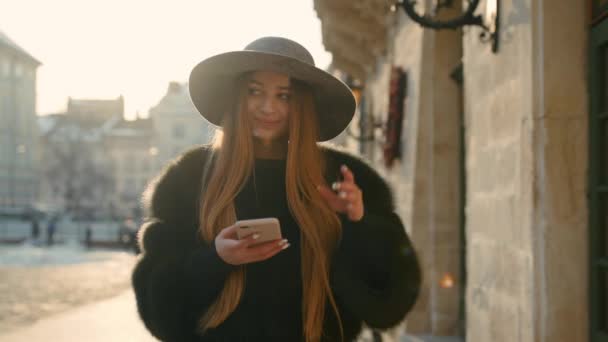 Superbe femme brune dans un chapeau gris debout dans la rue bondée de la ville et utilisant son téléphone pour envoyer un message texte — Video