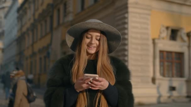 Mujer con un sombrero gris de pie en la concurrida calle de la ciudad y usando su teléfono para enviar mensajes de texto — Vídeo de stock