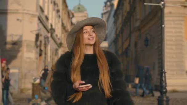 Mujer joven en un sombrero gris y pelo largo se comunica a través de teléfono inteligente — Vídeo de stock
