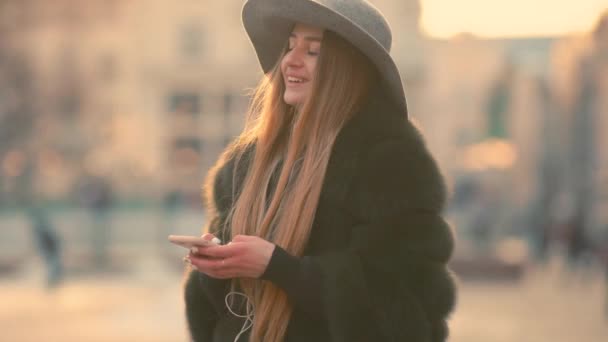 Hermosa mujer morena en un sombrero gris de pie en la concurrida calle de la ciudad y usando su teléfono — Vídeo de stock